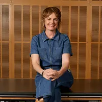 A woman sits on a black wooden bench smiling. She is wearing a denim jumpsuit, with her legs crossed and her hands resting on her knees.