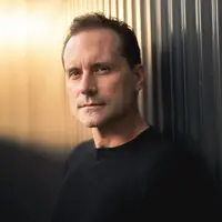 A man glares to the distance. He is wearing a black t-shirt and stands against a grey backdrop.