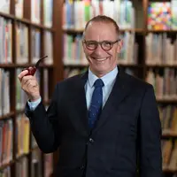 A man is standing in front of shelves of books. He is wearing glasses, a suit and tie and is holding up a pipe in his right hand.
