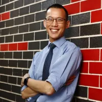 A man wearing glasses, a tie and buttoned blue shirt is standing against a wall smiling.