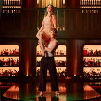 A man in black pants and a white tank top lifts a woman in a lacy outfit above his shoulders.