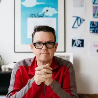 Geoff Cobham wearing a red t-shirt and black framed glasses, sitting at an office desk.
