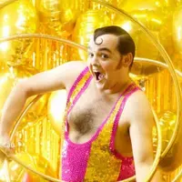 A man stands on an angle, wearing a sequined yellow and pink unitard with multiple hula hoops