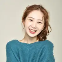 A woman with reddish brown hair stands in a blue jumper smiling.