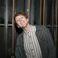 Josh Thomas wearing a striped shirt and checked over-shirt, smiling and leaning against a window.