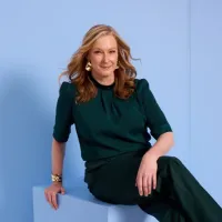 A blonde women wearing a green top and pants sits on top of a blue box leaning backwards resting on her arm.