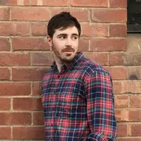 A bearded man wearing a read flannelette shirt stands against a brick wall.