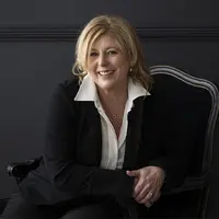 A woman with blonde hair sits in a regal looking black chair. She is wearing a black blazer and trousers and a white shirt with a wide collar.