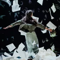 A women dances surrounded by falling paper. She is holding a book and her hair is in braids.