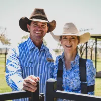 A man and woman stand smiling on a farm. Both are wearing wide brimmed hats and blue button shirts. The woman is wearing an arm brace.