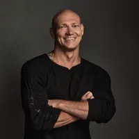 A bald man stands smiling with his arms crossed. He is in a long sleeve black shirt against a dark coloured background.