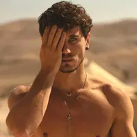 A man with brown curly hair stands close, one hand covering half his face, looking intensely into the camera.