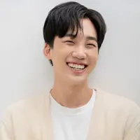 A man wearing a cream cardigan and white t-shirt stands looking off to his side smiling widely.