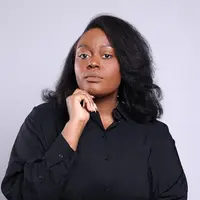 Professional studio portrait of a person with shoulder-length black hair wearing a black button-up shirt, resting their chin on one hand.