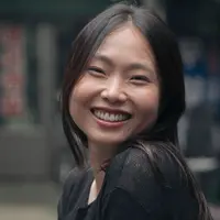 A woman is visible from the shoulders up. She is looking over her shoulder smiling wearing a black shirt.