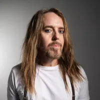 A man with long hair and a beard looks forward wearing a white t-shirt.