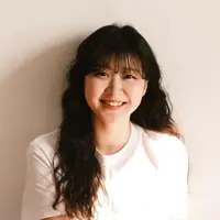 A woman in a light coloured shirt is wearing her long black hair down. She is smiling against a white backdrop.
