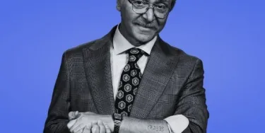 An older gentleman in in black and white against a blue background. He is wearing a suit and glasses and has rolled up his sleeve to display a tattoo.