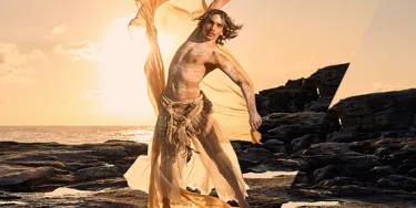 A dancer stands on a rock shelf in front of the ocean. It is sunrise and they are wearing a light yellow fabric that is flowing through the air.