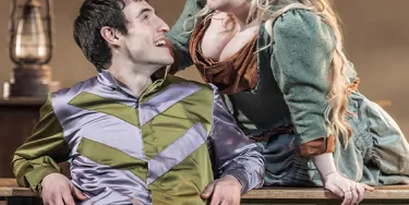 Two actors are seen on the set of the National Theatre Live production The Playboy of the Western World. A man sits leaning back on a desk while a woman leans over him from behind the desk.