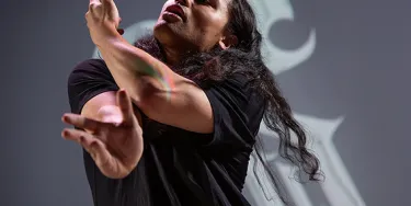 A dancer with long black hair, wearing a black t-shirt is looking upwards with their eyes closed. They are moving their arms in front of them.