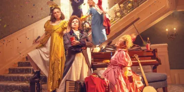 Five women stand staggered on a staircase in regency era gowns, with musical instruments for Pride and Prejudice (sort of) as confetti falls around them.