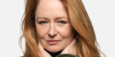 A woman with red hair wearing a green sweater holds her hands towards her neck.