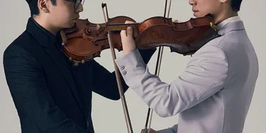 Two Asian men in their 30s face each other, both holding violins. One wears a black suit, the other a white suit.