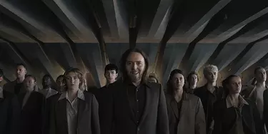 Tim Minchin, a man with long hair and a beard, stands in front of a group of people in a tunnel under the Opera House steps