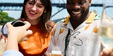 A man and woman stand smiling with glasses of champagne in their hands. The Harbour Bridge is visible in the background.