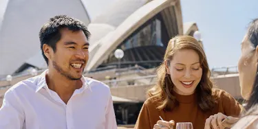 Three people sit around a table in the foreground of the Opera House sails. They are eating food and have drinks in front of them.