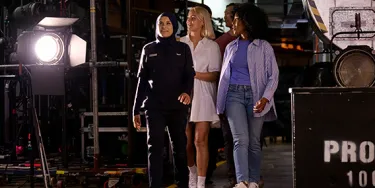 A group of people walk through a backstage area. There are lights and other production equipment surrounding them.