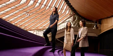 Three people climb the purple carpeted steps of the Opera House foyer.