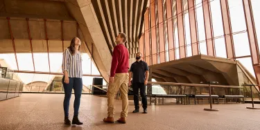 A man and woman stand next to a tour guide inside the Sydney Opera House foyer. They are looking up at the expansive windows and high ceilings.