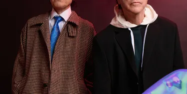 The two members of sparks stand facing the camera in front of a red background.