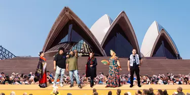Three men and three women from different backgrounds stand on a yellow platform in front of the Sydney Opera House sails in front of an audience.
