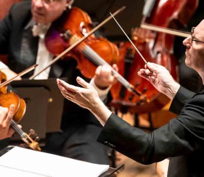 A conductor leads an orchestra as musicians play string instruments, including violins and cellos.