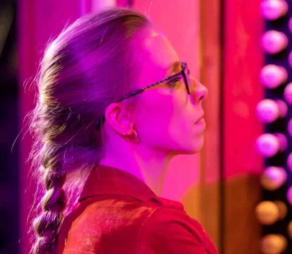A woman with glasses and a long braid, wearing a red shirt, is illuminated by pink and purple light while playing a pipe organ.