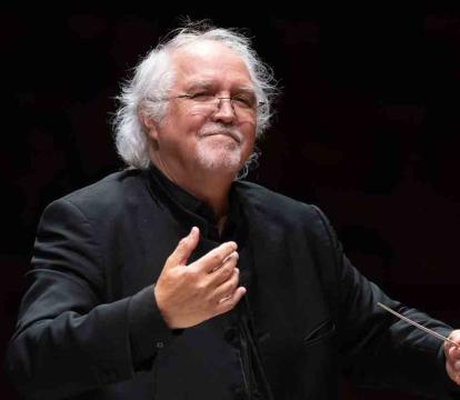 An older man with white hair, wearing glasses and a black suit, stands conducting with a baton in his right hand.