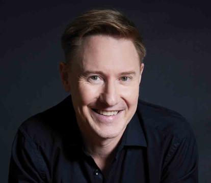 A smiling man with light brown hair wearing a dark shirt sits against a dark, softly lit background.