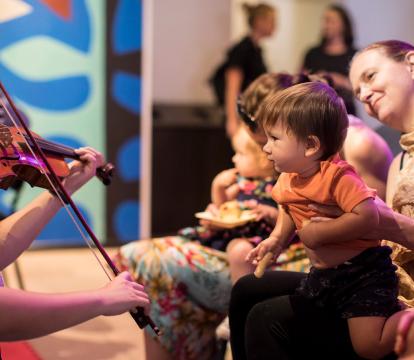 A man with grey hair and a beard plays a violin which is resting on his lap.