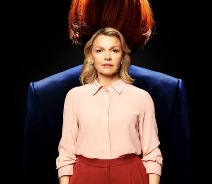 A white woman with shoulder-length blonde hair wears a pink shirt and red pants, looking at the camera.