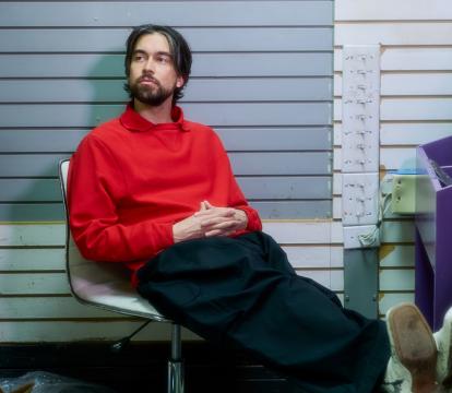A white man sits in a chair with his feet up wearing a red sweater.