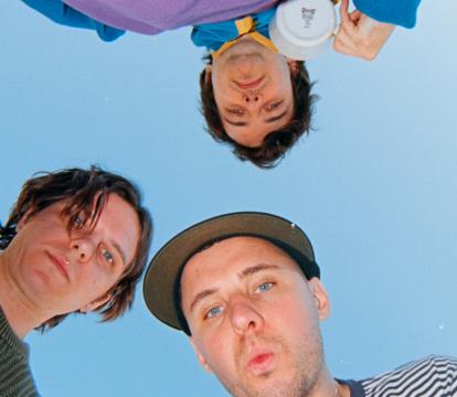 Three people look down at the camera, faces curious and playful. They are outside under a clear blue sky.
