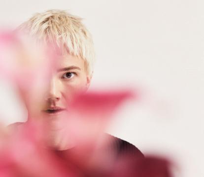 A person with short blonde hair standing behind some pink foliage against a white background.