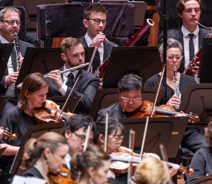 An orchestra performing. We see men and women playing a variety of instruments including violins, oboes, trumpets and flutes.