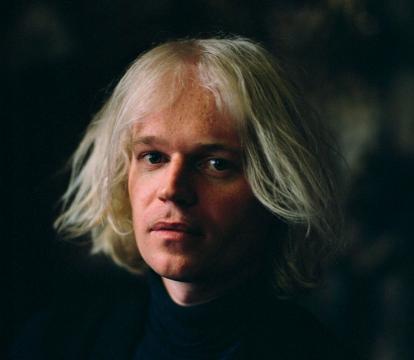 A white man in early 40s with jaw-length white hair looks at the camera.