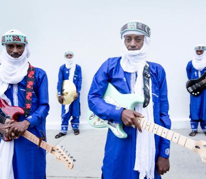 Four men wearing blue outfits and white headscarves, three holding a guitar and one holding a drum cymbal.