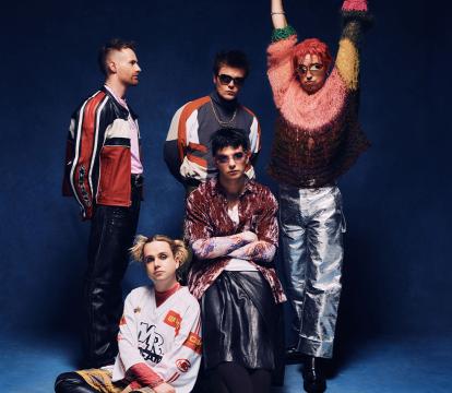 A group of five men dressed in kooky, punky Y2K outfits pose in front of a blue backdrop.