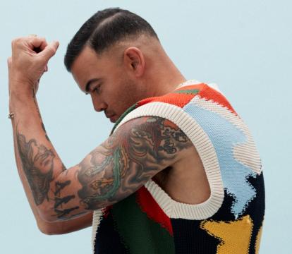 A man with tanned skin and short black hair is posed standing on his toes, with bent knees and his arms held up in front of his face. He wears maroon pants, green and yellow Nike shows and a multi coloured vest.